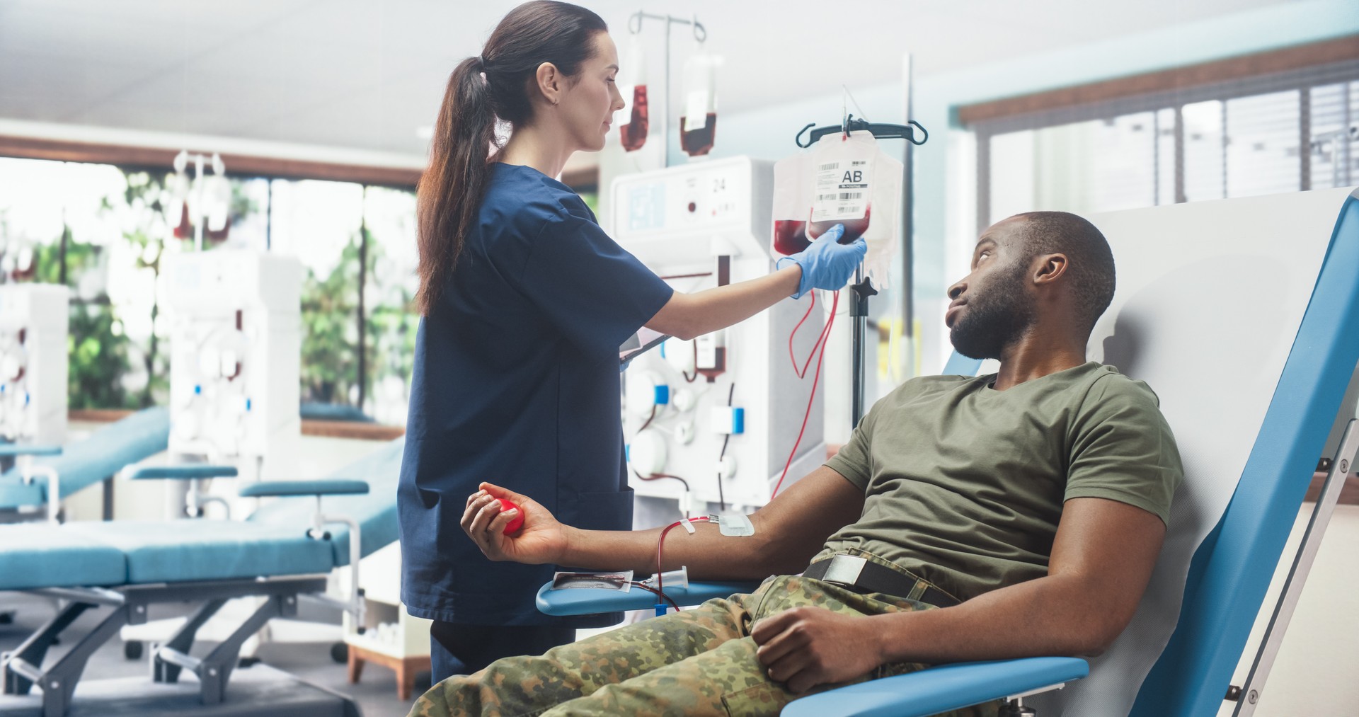 Caucasian Female Nurse Taking Blood Donation From Black Army Troop In Military Hospital. African American Man Squeezing Heart-Shaped Ball To Pump Blood Into Bag. Soldier Donating To Wounded Comrades.