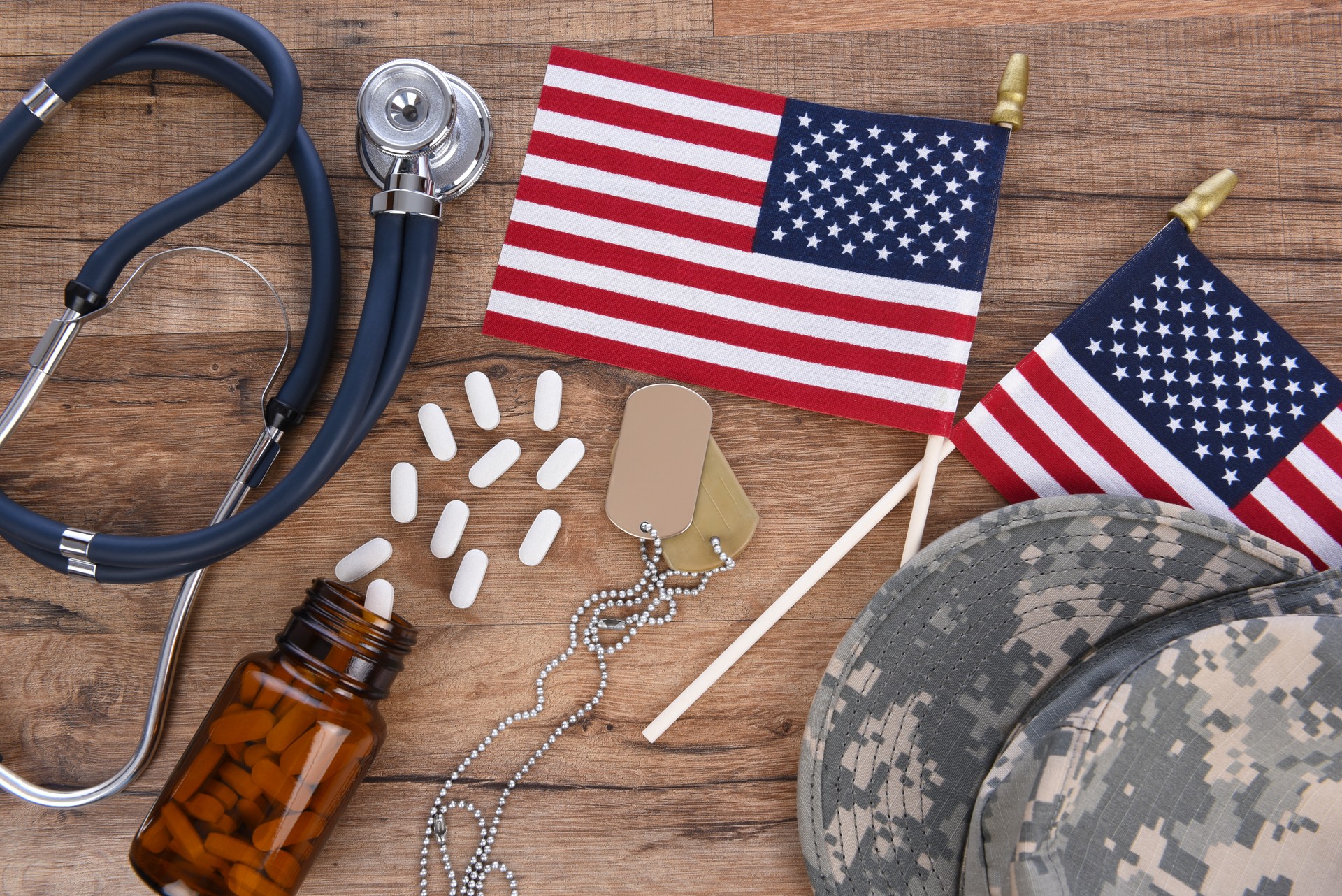 Military and Veterans Health Care Concept. Camo hat, dog tags, stethoscope, pills, and American Flags on a wood background.